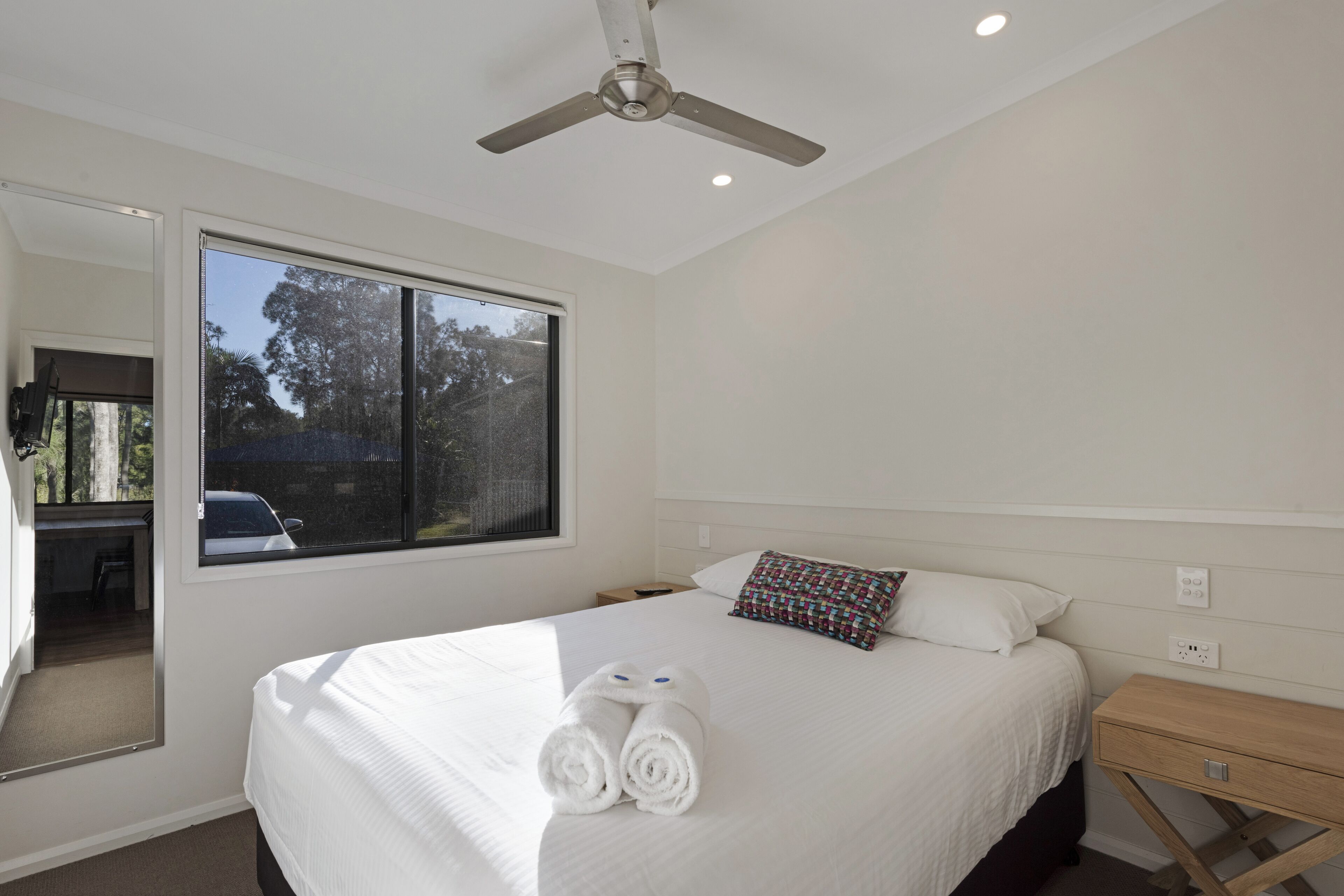A neatly made bed with rolled towels, a bedside table, a nightstand, a ceiling fan, and a window with a view of trees and a car outside.