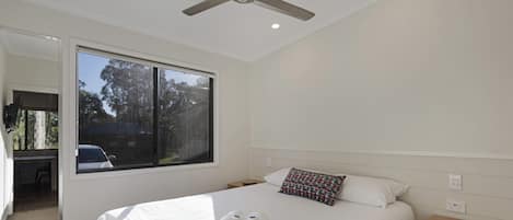 Un lit bien fait, avec des serviettes pliées, une table de chevet, une commode, un ventilateur de plafond et une fenêtre donnant sur des arbres et une voiture garée à l’extérieur.