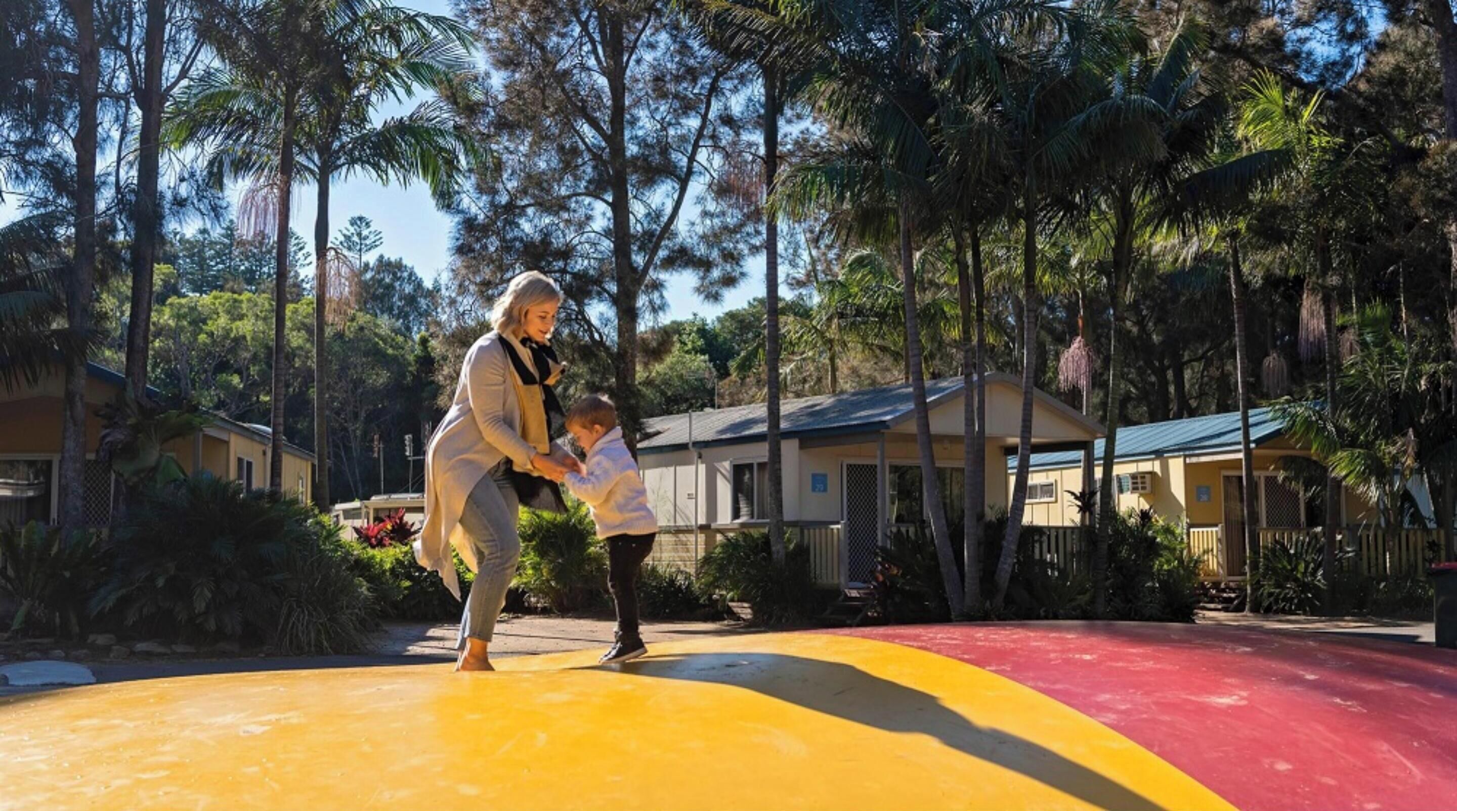 Une femme et un enfant jouent sur une aire de jeux colorée, avec des palmiers et des bâtiments en arrière-plan.