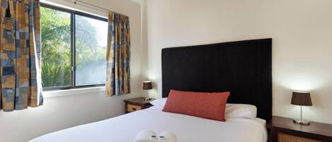 Habitación de hotel con una cama grande, una ventana con cortinas, una mesita de noche con lámpara y vistas a zonas verdes.