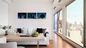 A modern living room with a sofa, a dining table with a book and a bowl of fruit, and a city view through large windows. - The Putman (Hong Kong)
