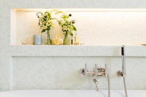 A bathroom with a white bathtub, a shower with a handheld showerhead, and a shelf with toiletries and a floral arrangement.