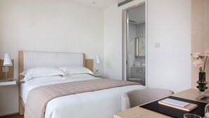 A hotel room with a bed, bedside table, lamp, chair, and a desk with a book and glass. - The Putman (Hong Kong)