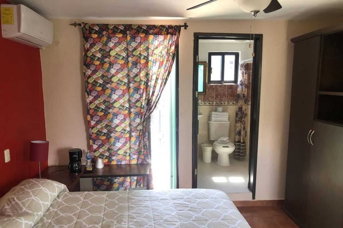 A bedroom with a bed, a desk, a chair, a wardrobe, and a bathroom visible through an open door.
