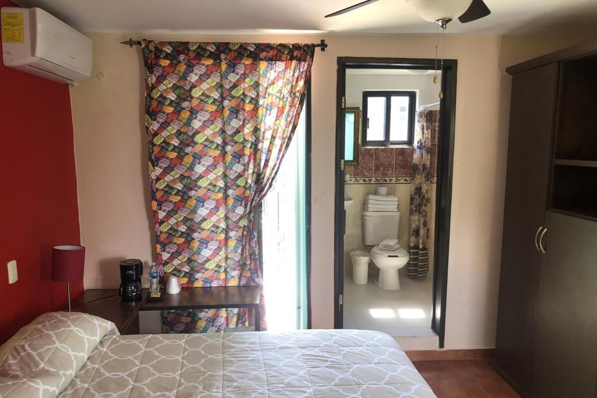 Une chambre à coucher comprenant un lit, un bureau, une chaise, une armoire et une salle de bain visible à travers une porte ouverte.