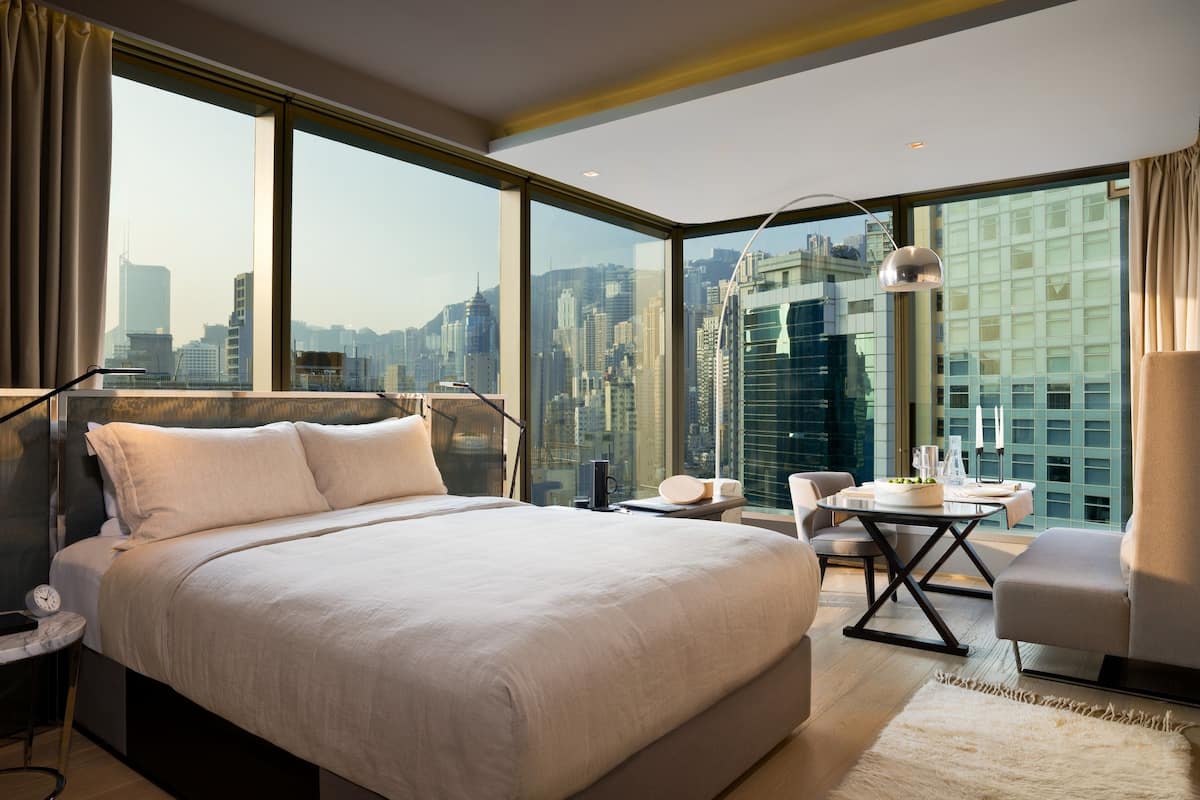 A modern hotel room with a large bed, a desk, a chair, and a view of the city skyline through large windows.