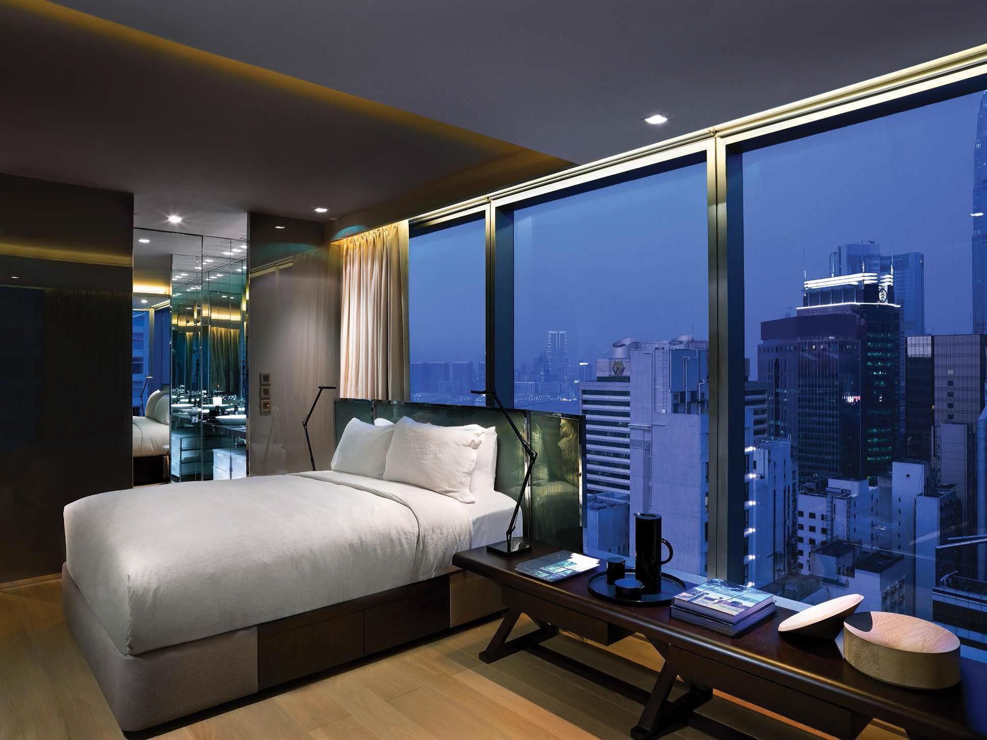 A modern hotel room with a large bed, a desk with a computer, and a view of a cityscape.