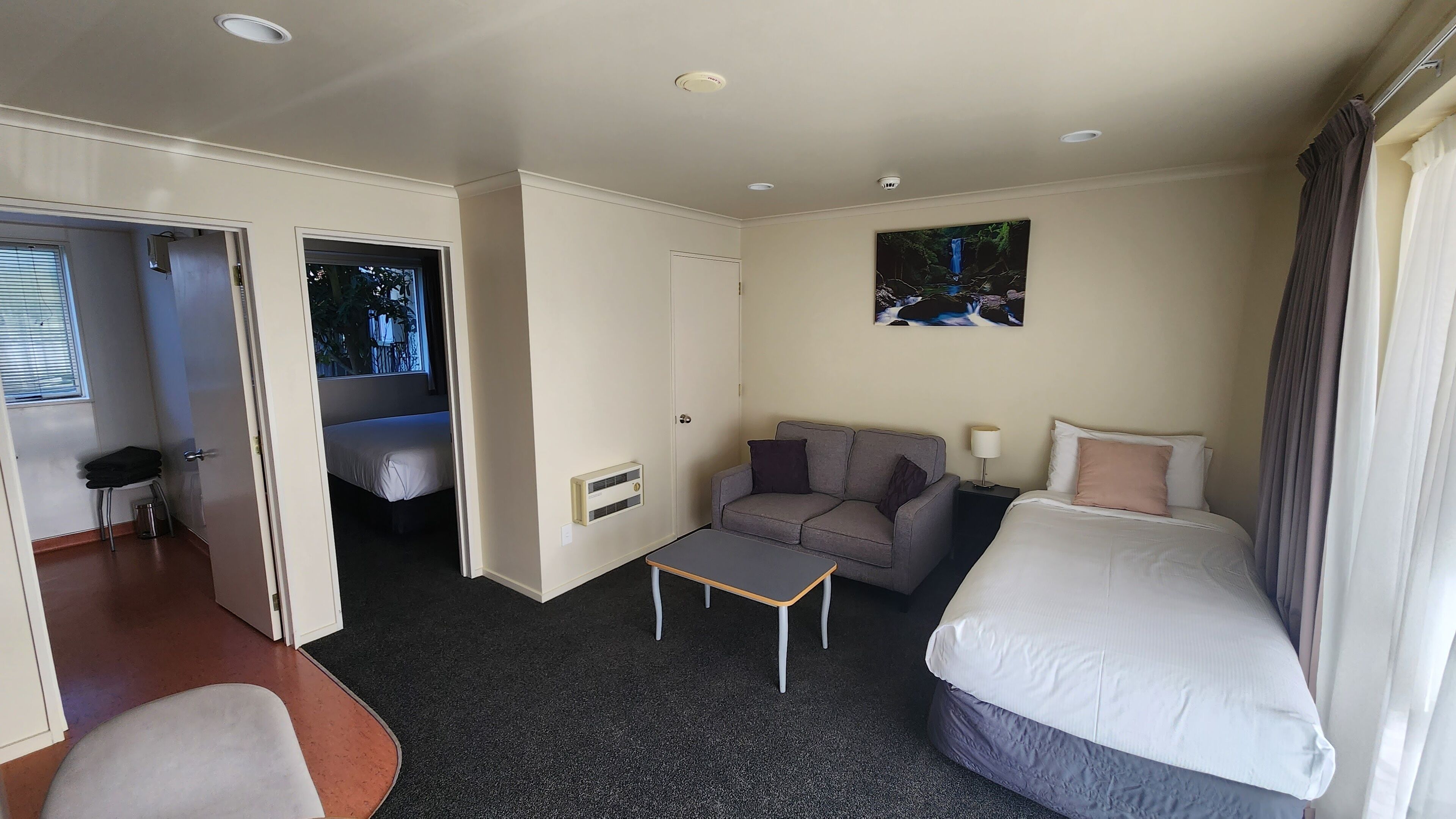 Een hotelkamer met een bed, een bank, een kleine tafel en een stoel. Er is een deur naar een balkon met uitzicht op bomen.