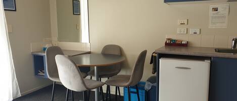 A compact kitchen with a refrigerator, a microwave, and a sink. There is a small dining table with four chairs. A flat-screen TV is mounted on the wall above the table.