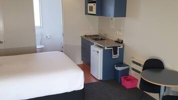 Een compacte hotelkamer met een eenpersoonsbed, een kleine kitchenette, een bureau en een stoel.