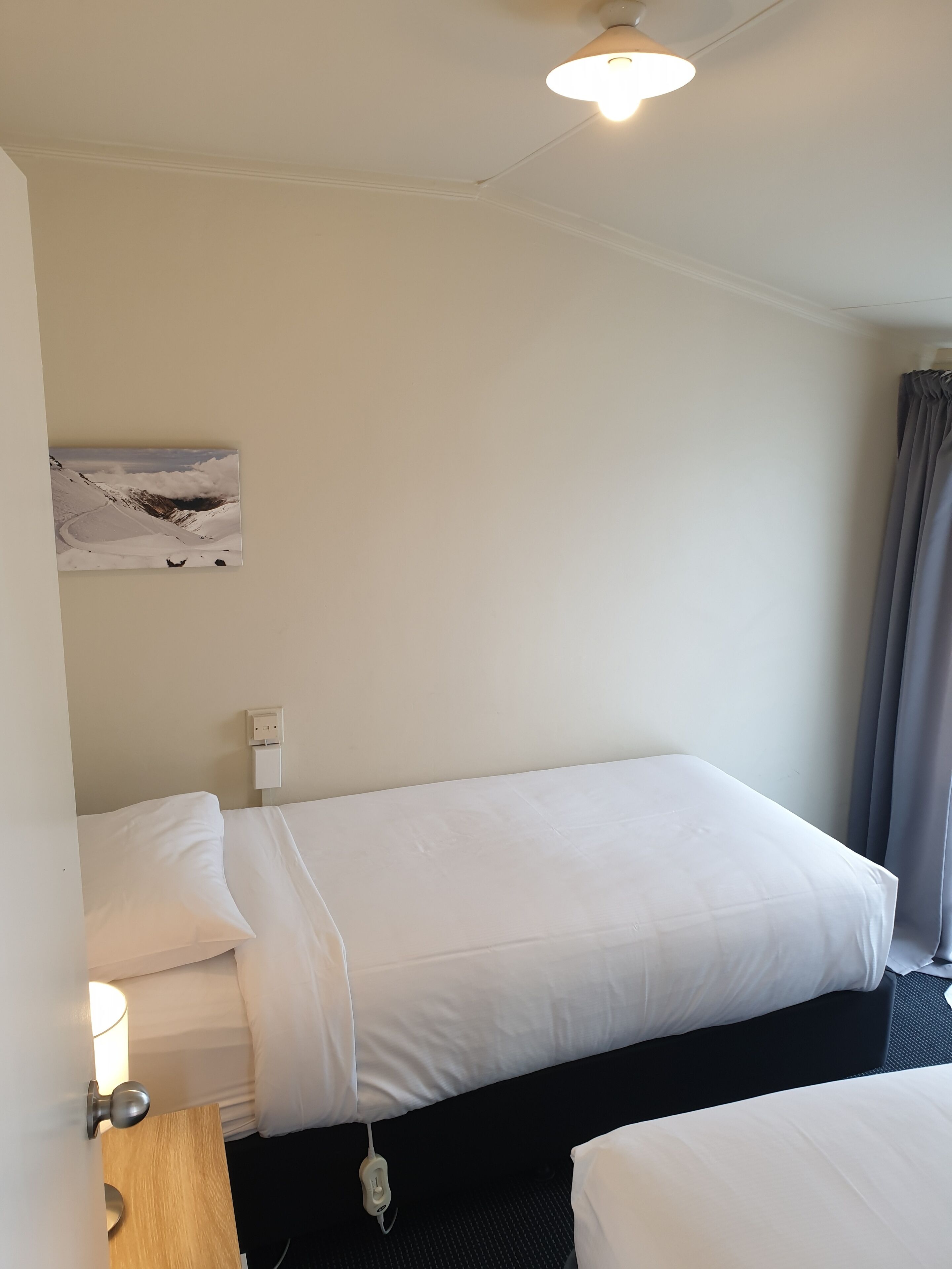 A hotel room with a single bed, a bedside table with a lamp, a power outlet, and a painting on the wall.
