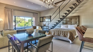 A modern bedroom with a large bed, a dining area with a glass table, and a staircase. - Heritage Square Apartments (Cape Town)