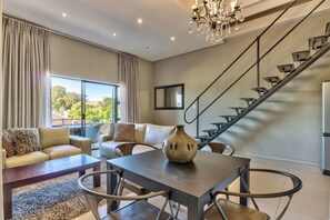 A modern living room with a sofa, coffee table, and a vase. There is a staircase with a railing and a chandelier. - Heritage Square Apartments (Cape Town)