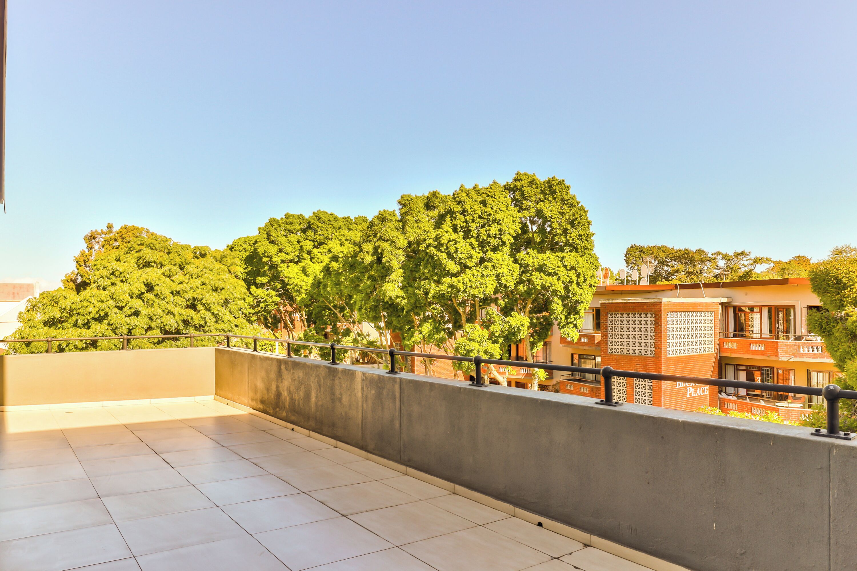 A rooftop terrace with a concrete floor, a metal railing, and a view of trees and buildings.