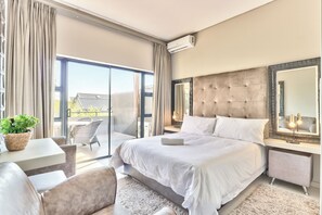 A modern bedroom with a large bed, a bedside table, a chair, a small table with a plant, and a balcony with a view. - Heritage Square Apartments (Cape Town)