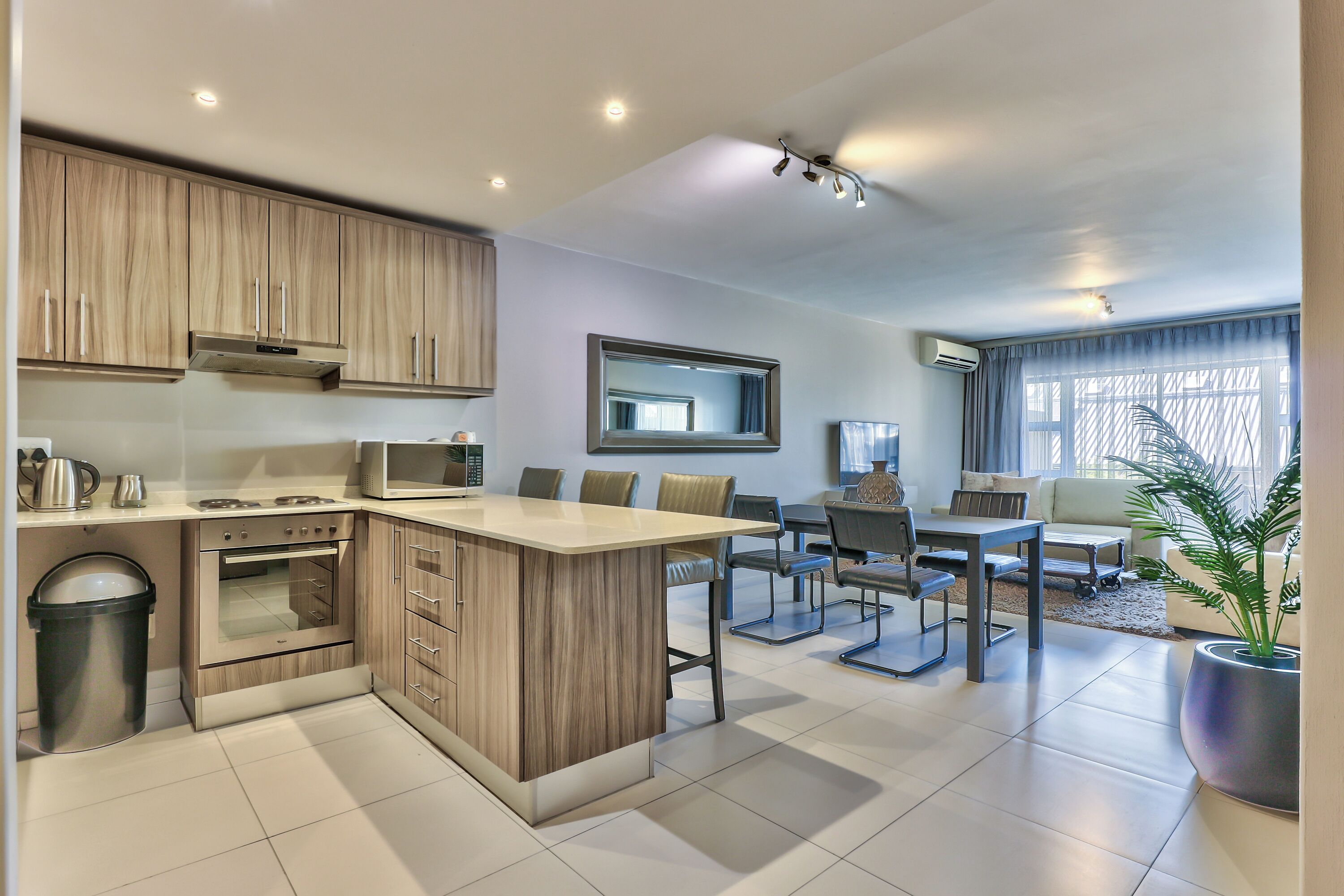 A modern kitchen with an island, built-in oven, and microwave. There is a dining area with a table and chairs, and a large window with curtains.