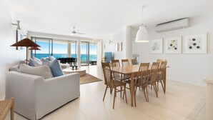 A spacious living area with a white sofa, dining table, and chairs, large windows with a view of the ocean, and ceiling fans. - La Mer Noosa (Noosa Heads)