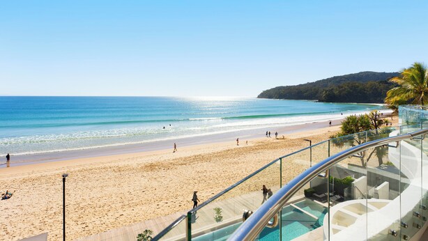 A beach view from a balcony with a glass railing, clear blue sky, and a sandy beach with people walking. - La Mer Noosa (Noosa Heads)