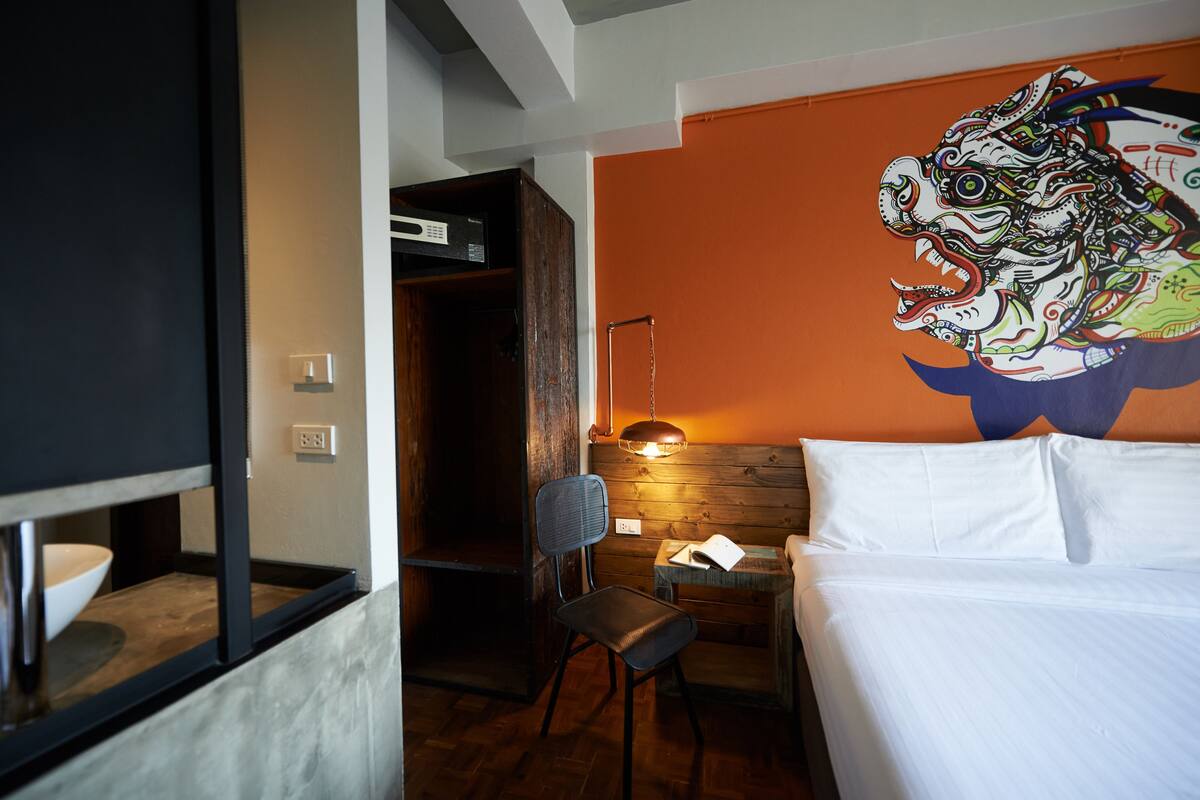 A hotel room with a bed, a chair, a desk with a lamp, and a wall mural.