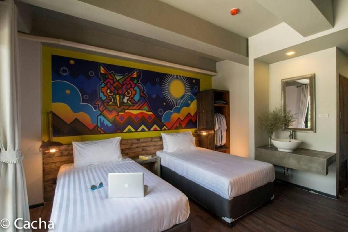 A hotel room with two beds, a laptop on one bed, a colorful mural on the wall, a wooden headboard, a bedside table, a nightstand, a mirror, a sink, and a plant.