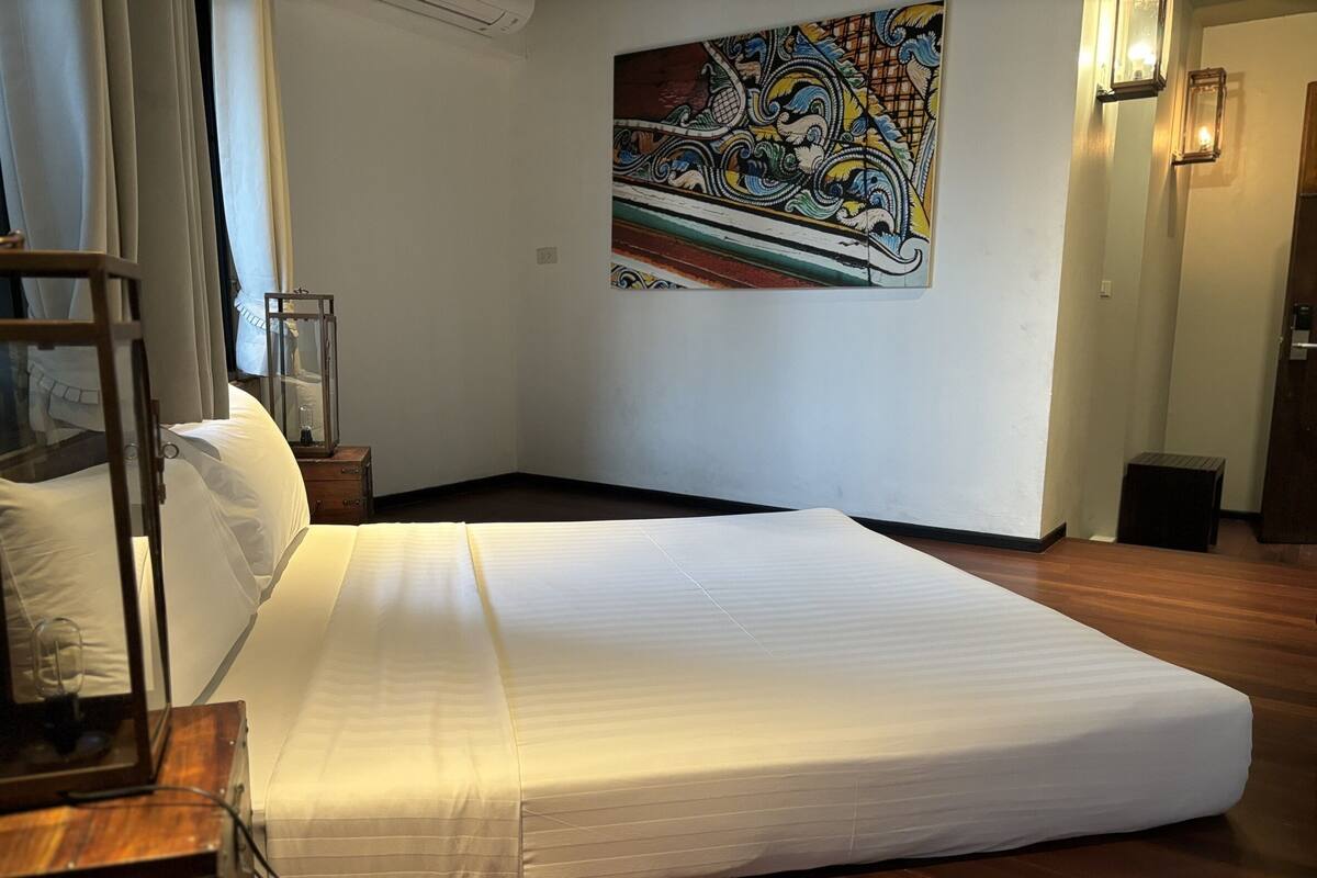 A room with a large bed, a wooden floor, a painting on the wall, and a small table with a lamp.