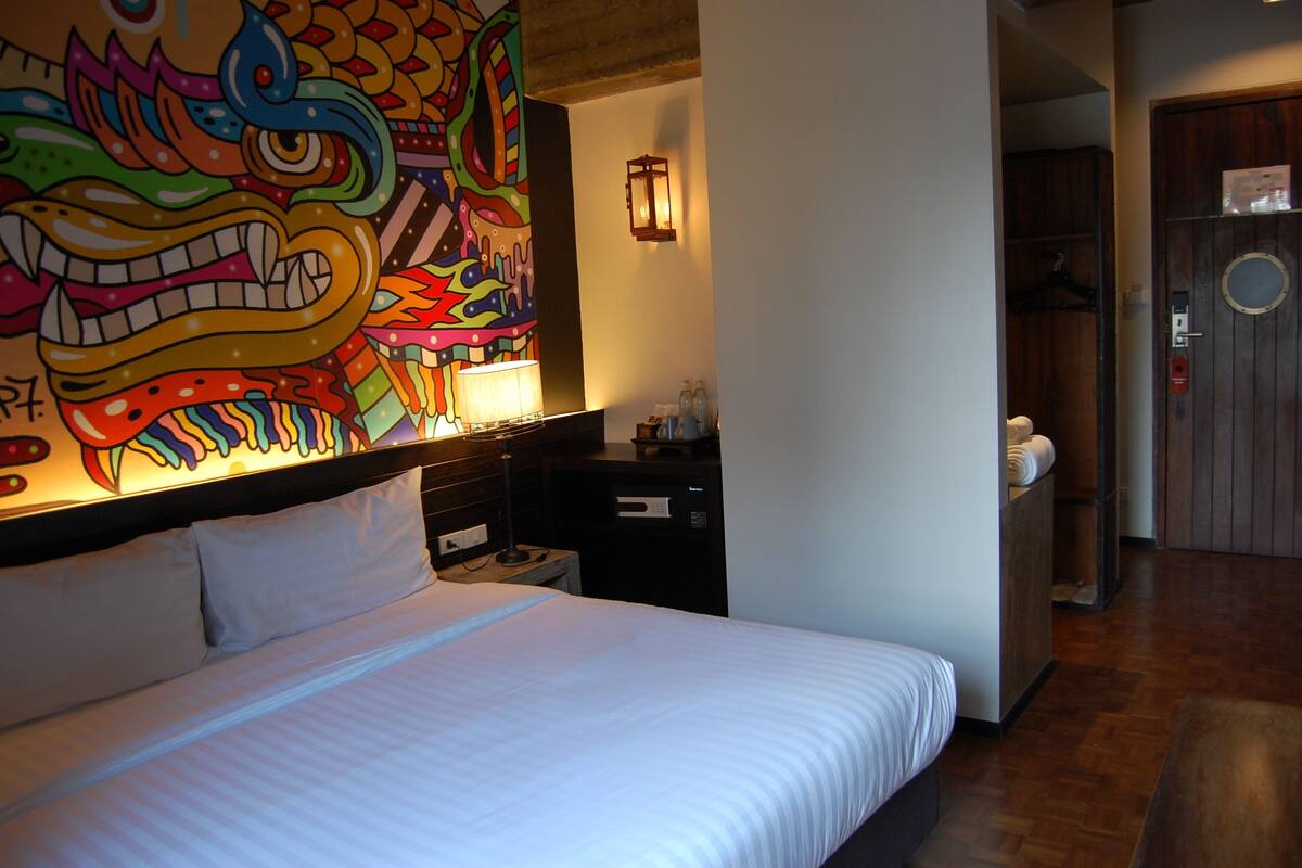 A hotel room with a large bed, a nightstand, a lamp, a wall with a colorful mural, a wooden door, and a small closet.