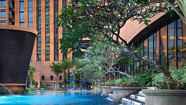 A hotel pool area with a modern building, a water feature, and lounge chairs.