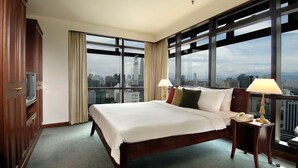 A hotel room with a large bed, two bedside lamps, a telephone, and a view of the cityscape through large windows.