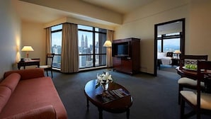 A hotel room with a large window offering a city view, a sofa, a coffee table with magazines, a TV, and a dining area with a table and chairs.
