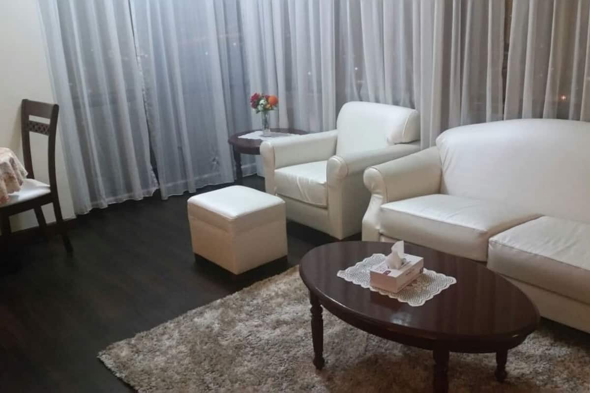 A living room with a dark wooden floor, a round coffee table with a tissue box, a beige sofa, a white armchair, and a small table with a vase of flowers.