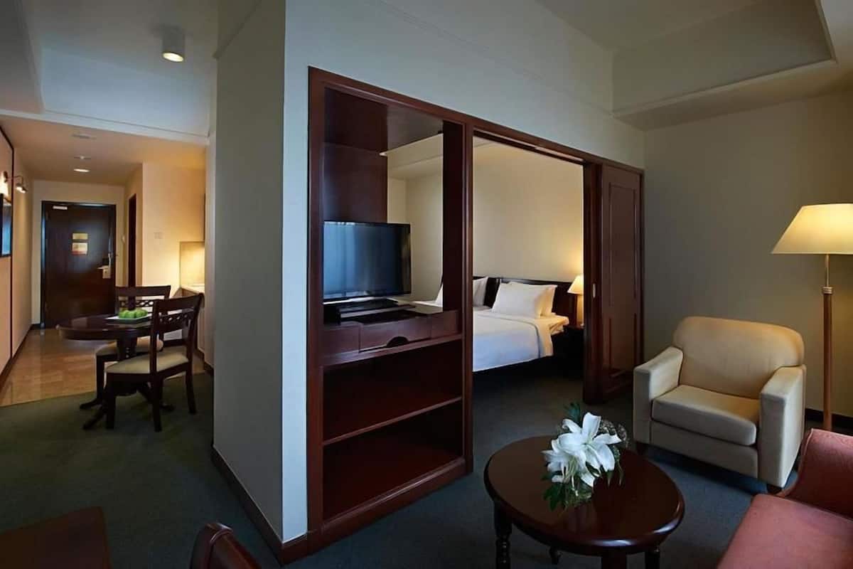 A hotel room with a bed, a desk with a TV, a chair, a small table with a flower, and a lamp.