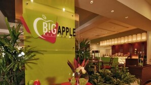 A reception area with a green wall displaying the Big Apple Restaurant sign, a red reception desk, and a variety of indoor plants.
