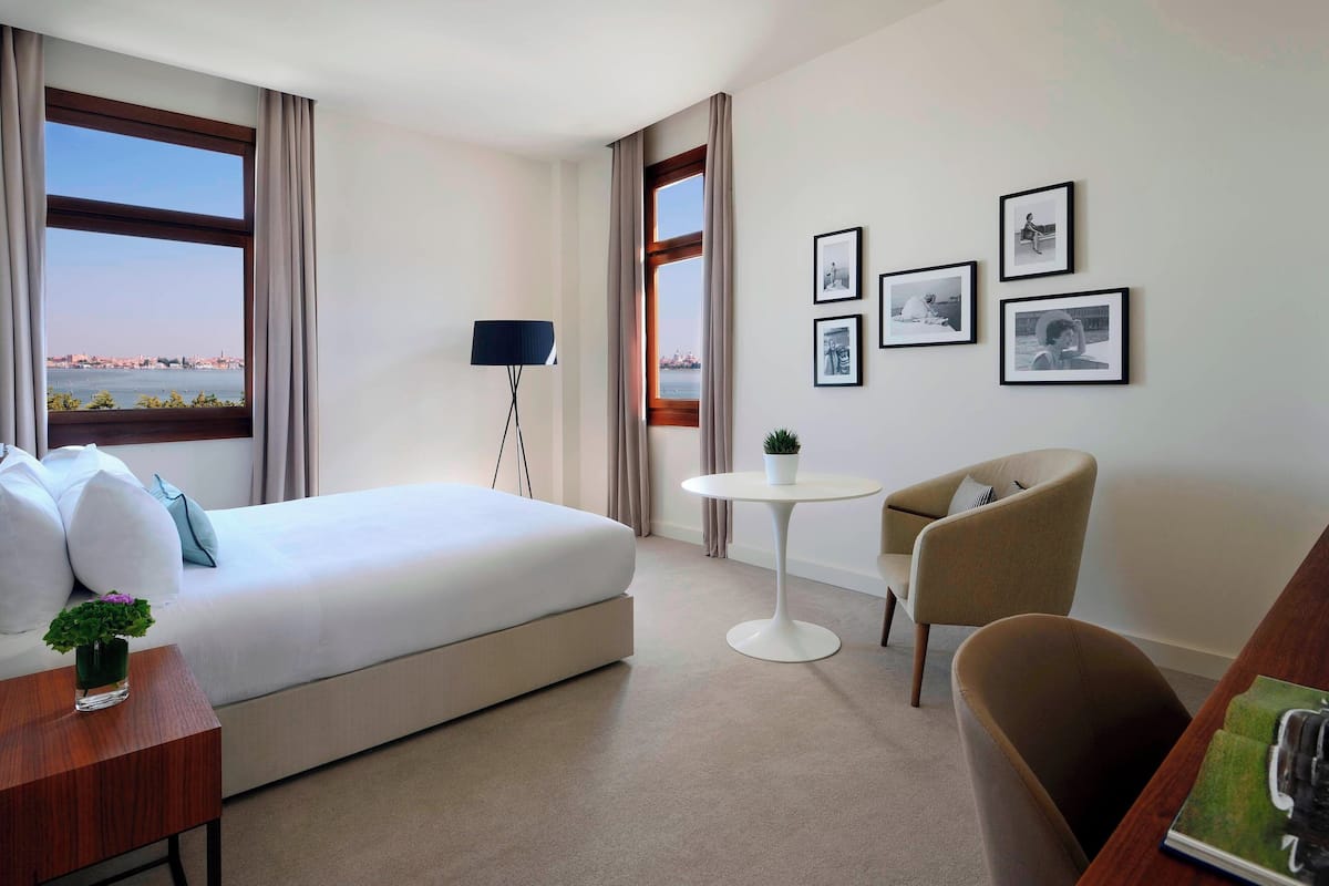A hotel room with a bed, a desk, a chair, a small table, and a view of a cityscape through large windows.