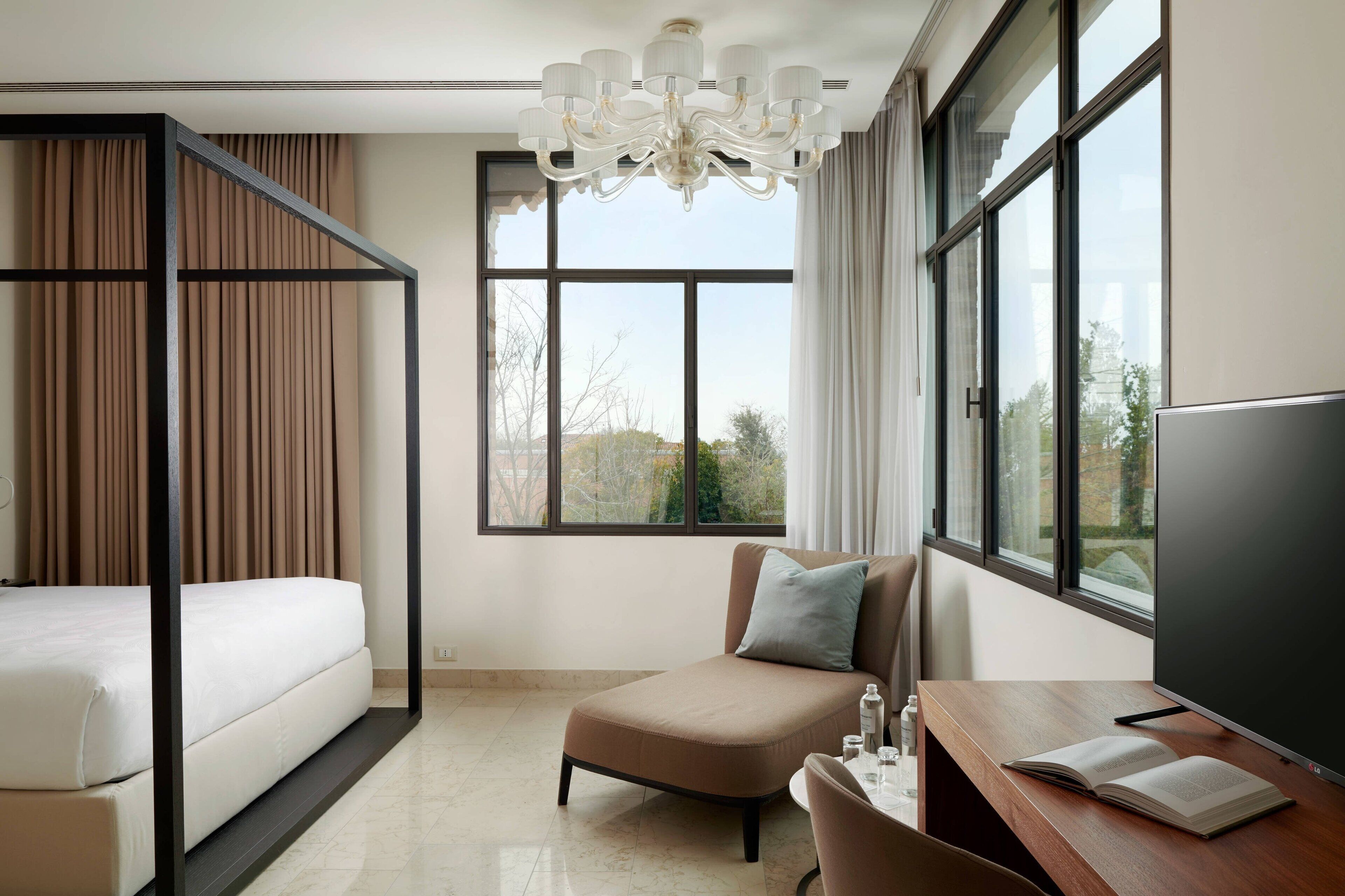 Quarto de hotel moderno com cama, sofá, escrivaninha com televisão e grandes janelas com vista para a cidade.