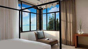 A modern hotel room with a large window offering a view of the sea, a bed, a seating area with a cushion, and a wooden side table with a vase.