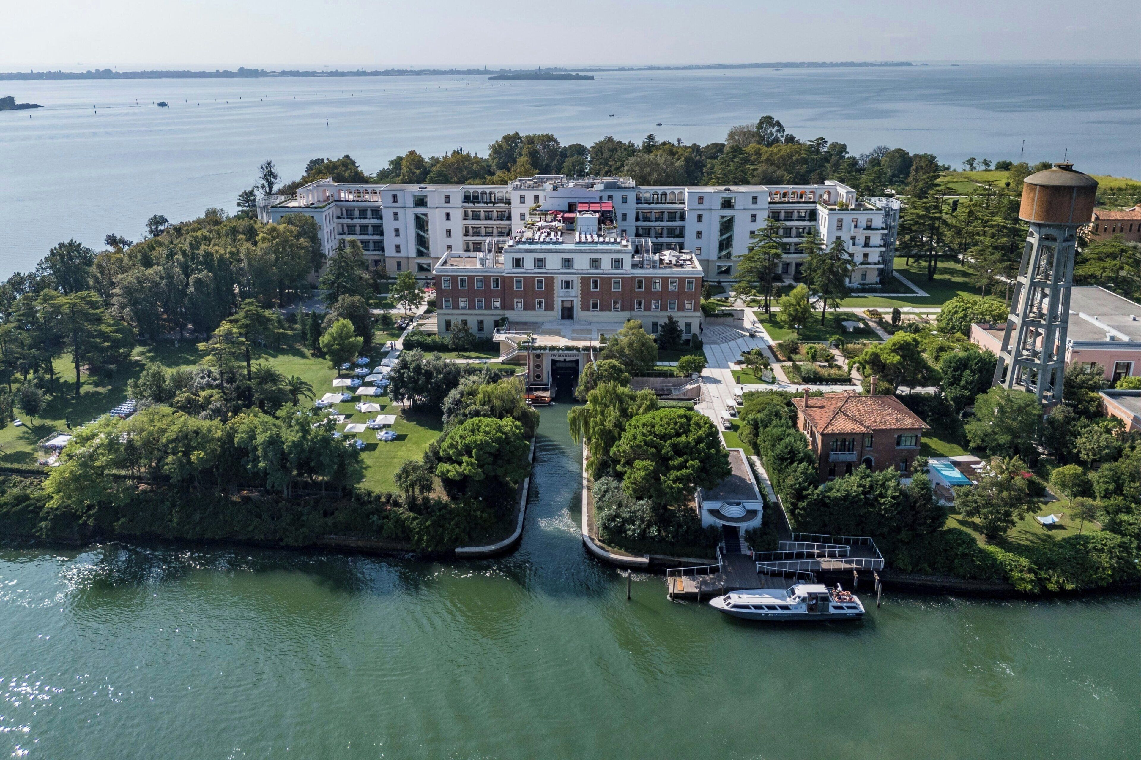 Vista aerea di un hotel sul lungomare, con un cortile centrale, una torre d'acqua e un molo con una barca.