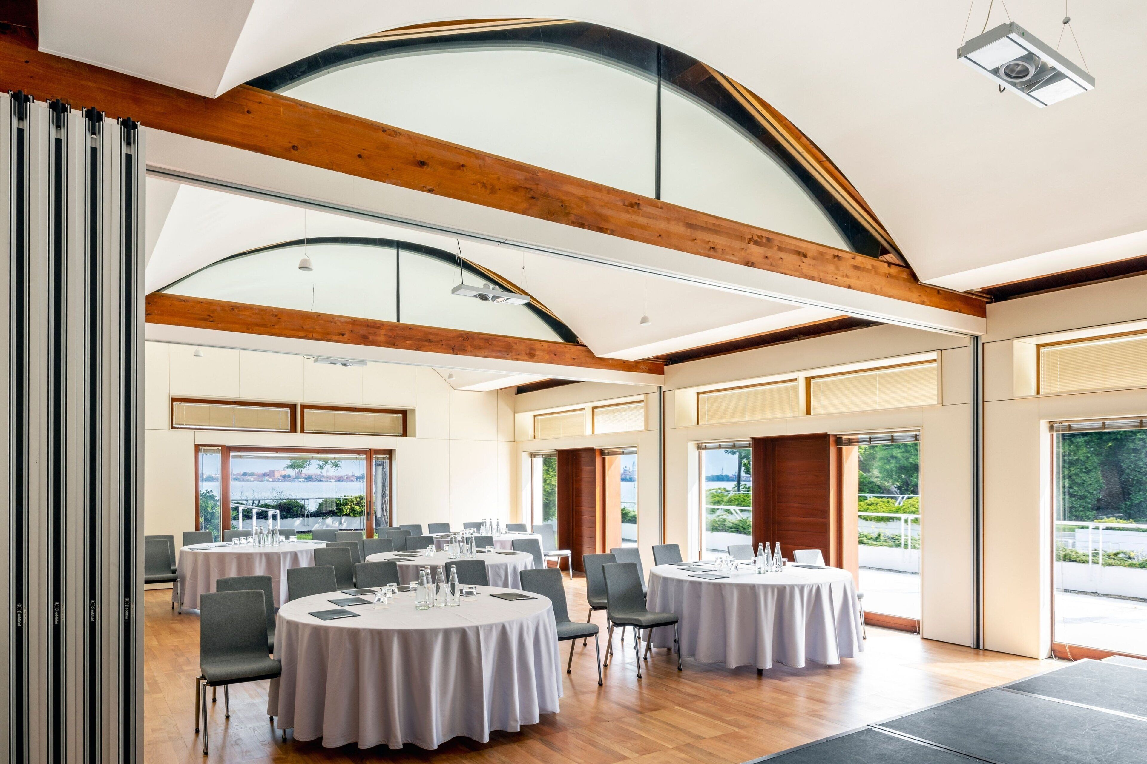 A spacious conference room with round tables, modern wooden beams, large windows, and a view of greenery.