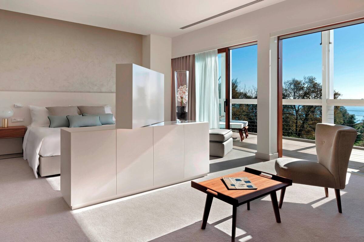 A modern hotel room with a bed, a sofa, a coffee table, and a view of the outdoors.