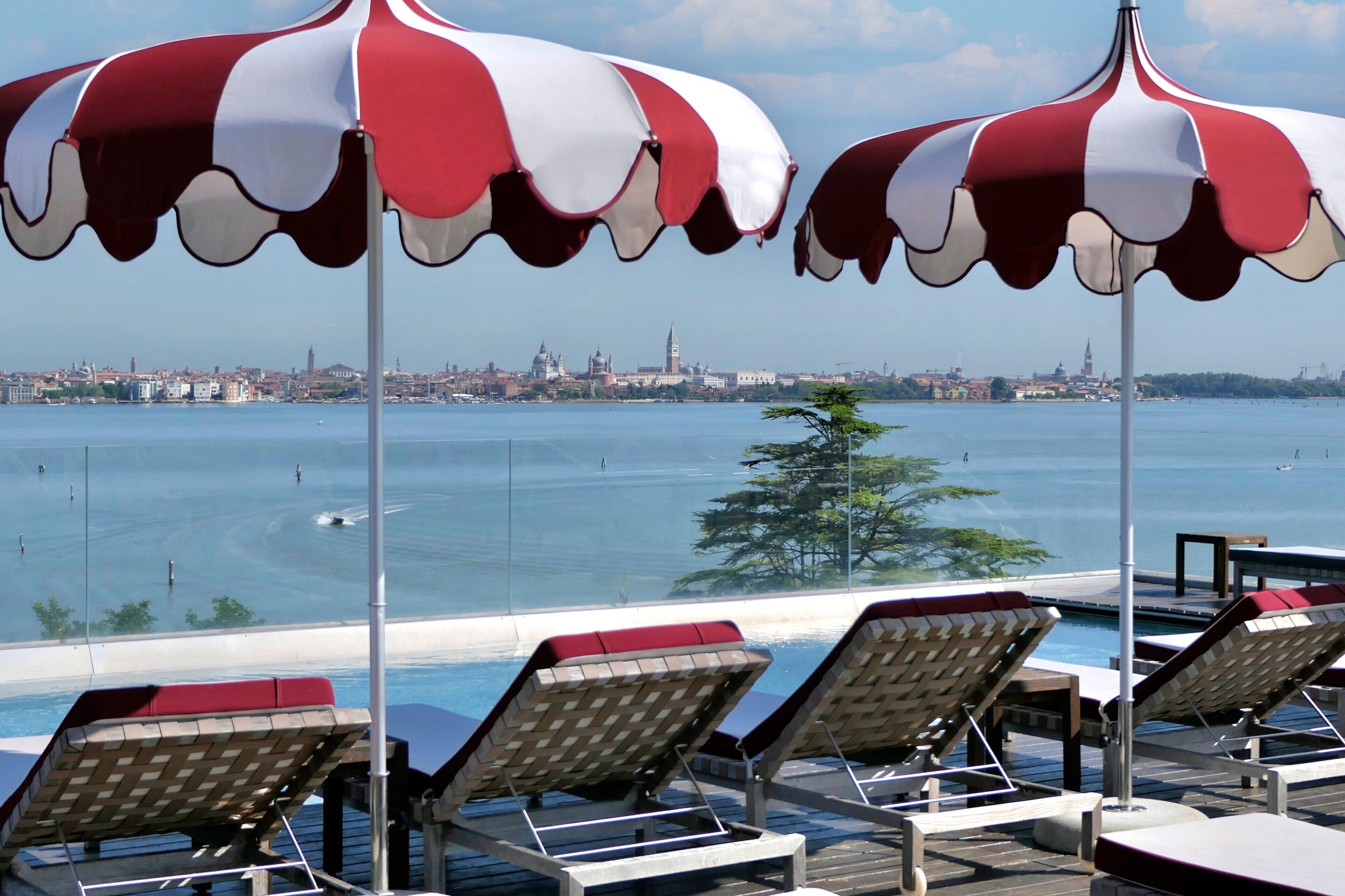 A poolside area with lounge chairs and red and white striped umbrellas, overlooking a body of water with a cityscape in the background.