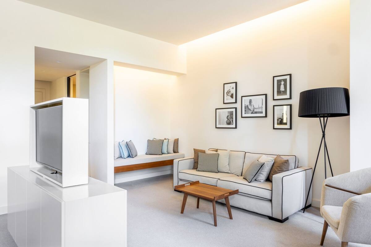 A modern living room with a white sofa, a wooden coffee table, and a TV on a white stand. There are framed pictures on the wall and a floor lamp.