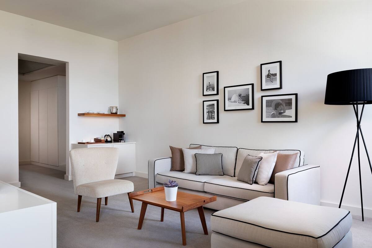 A modern living room with a sofa, armchair, and coffee table. There are framed pictures on the wall and a lamp on a tripod.