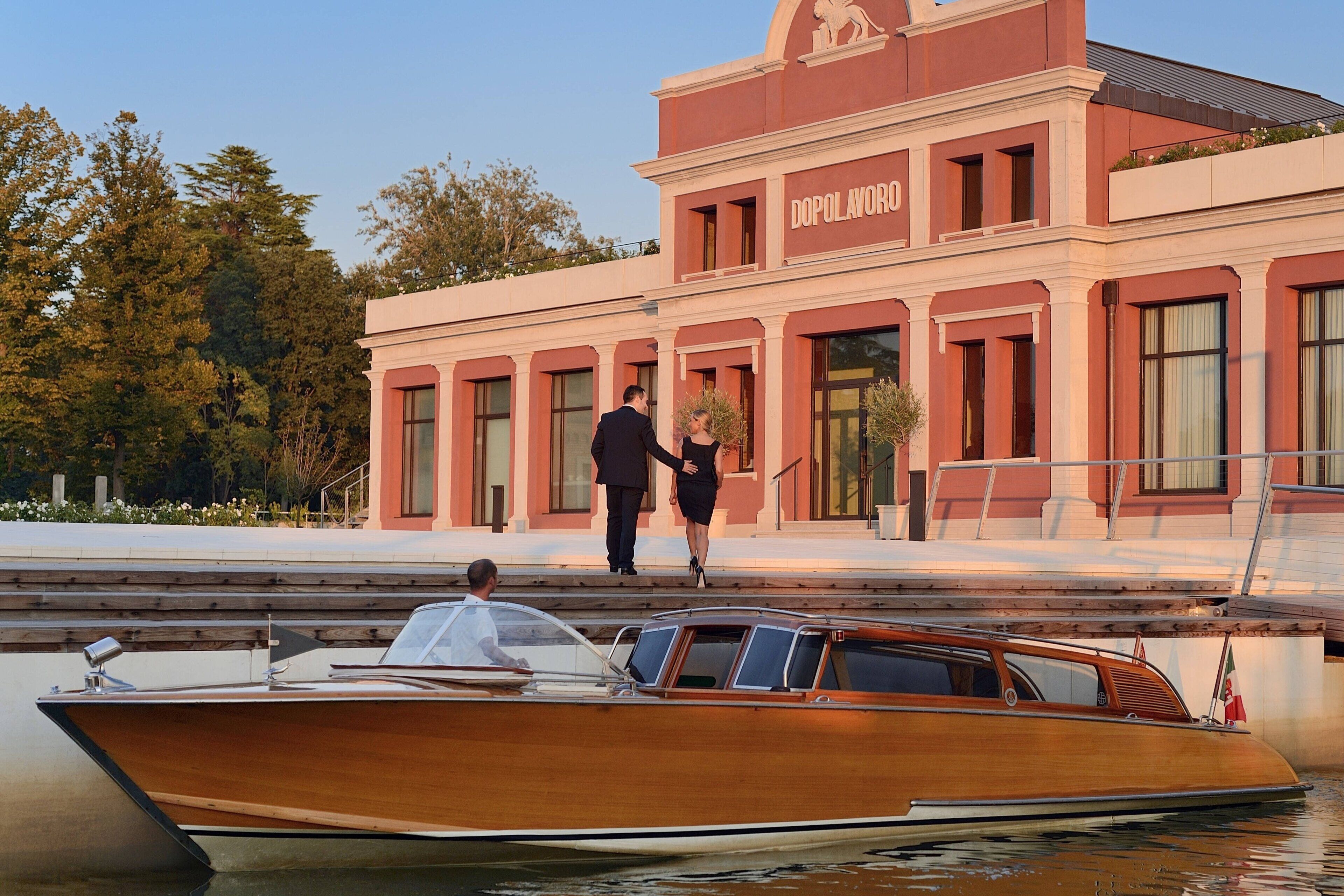 A wooden boat docked at a building with a red facade and the name "DOPOLAVORO" visible.