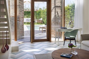 Una camera moderna con pavimento a spina di pesce, una scrivania con sedia, un tavolino da caffè con un libro e un vaso, e vista su un patio esterno con piscina e mobili.