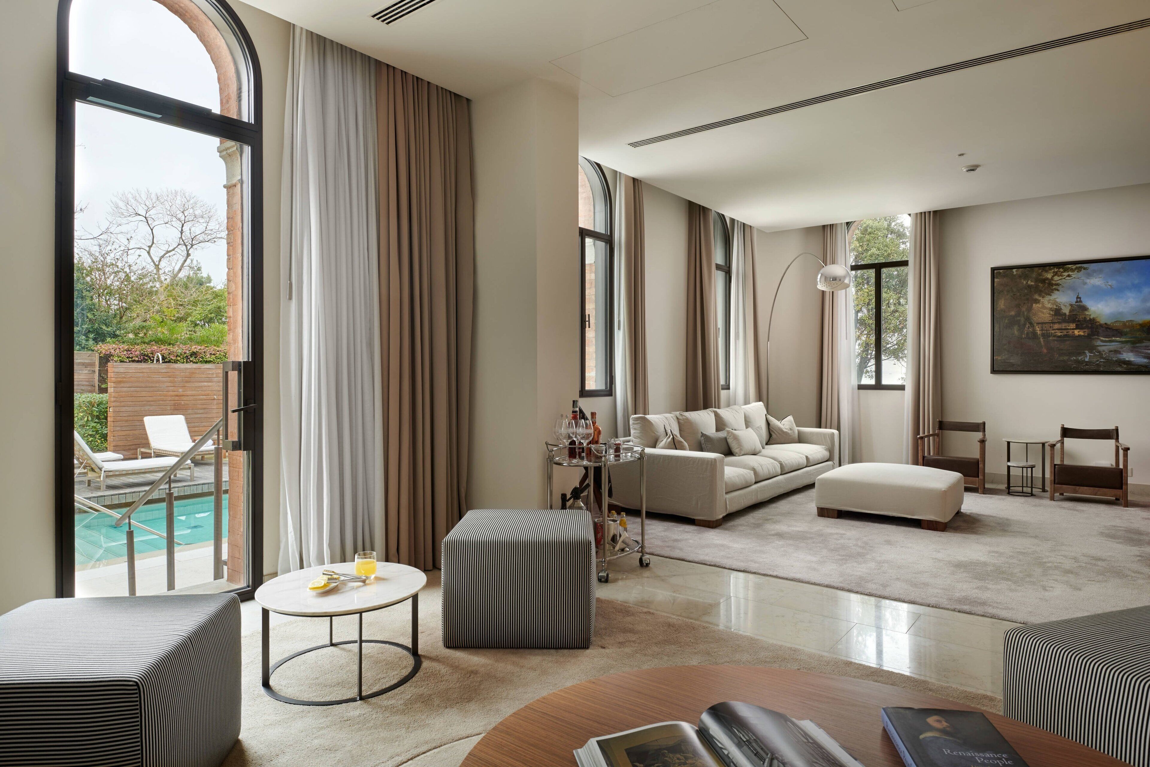 Amplia sala de estar con un gran ventanal, vista a la piscina, un sofá, otomanas y una mesa de centro.