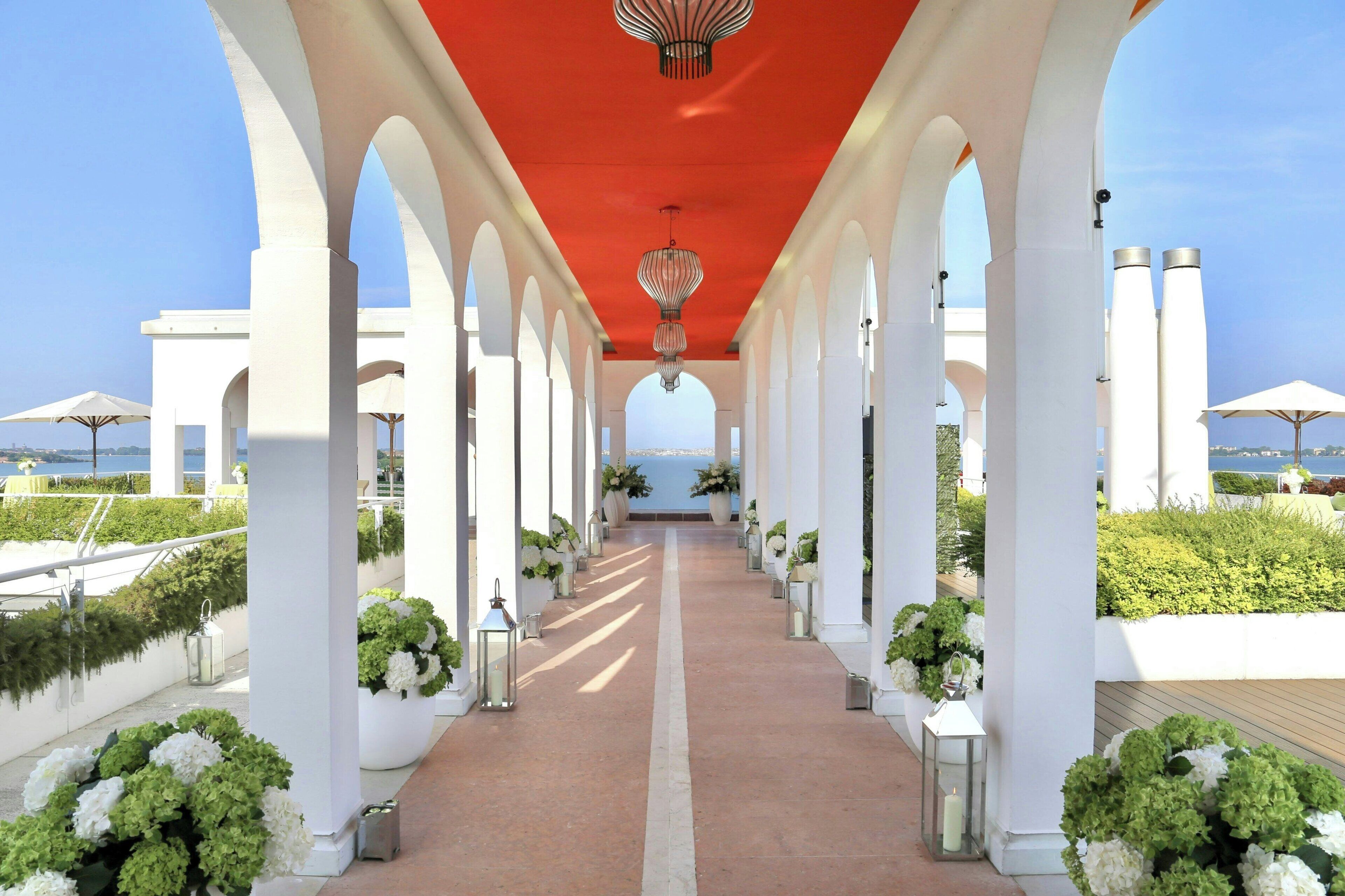 A corridor with white columns and a red ceiling, leading to a view of the sea.