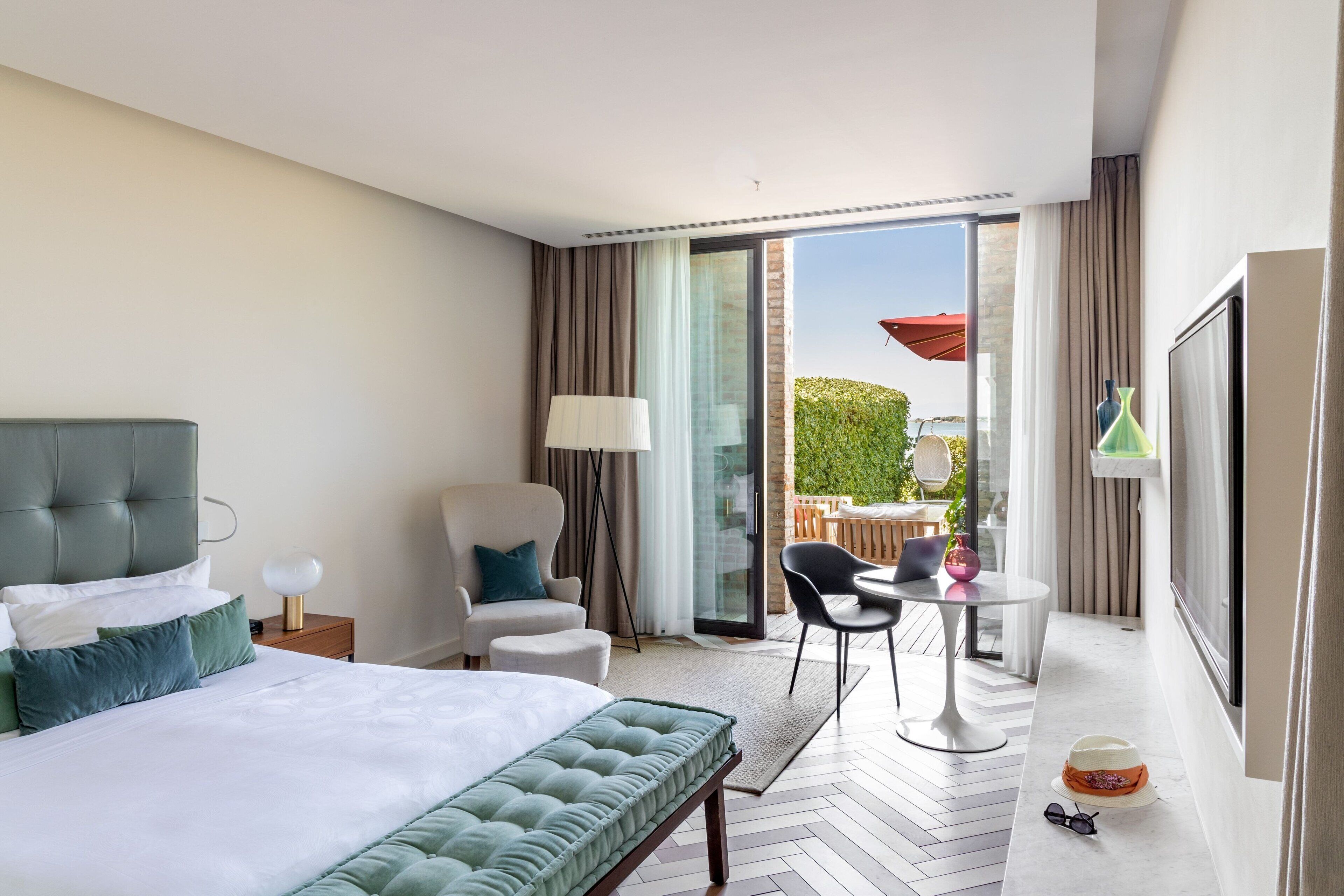 A hotel room with a large bed, a chair, a small table, and a balcony with a table and chairs.