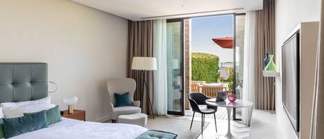 A hotel room with a large bed, a chair, a small table, and a balcony with a table and chairs.