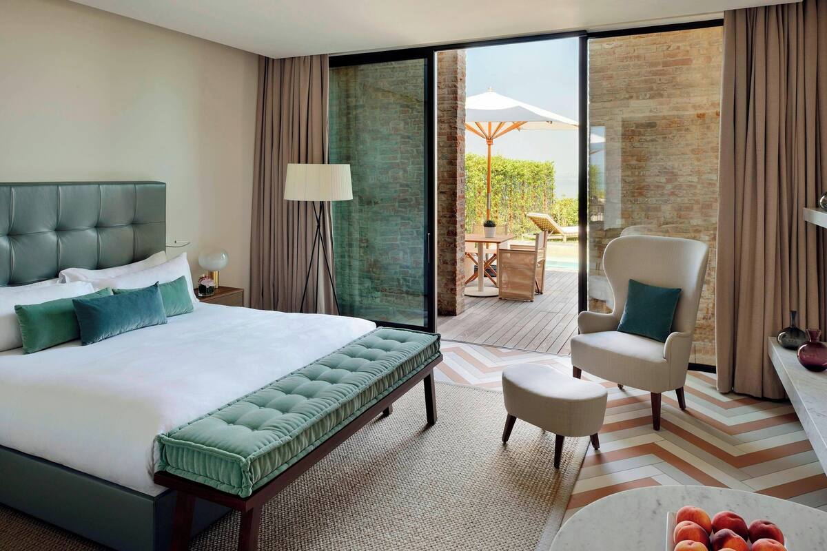 A hotel room with a large bed, a chair, a small table, and a view of an outdoor area with a table and chairs.