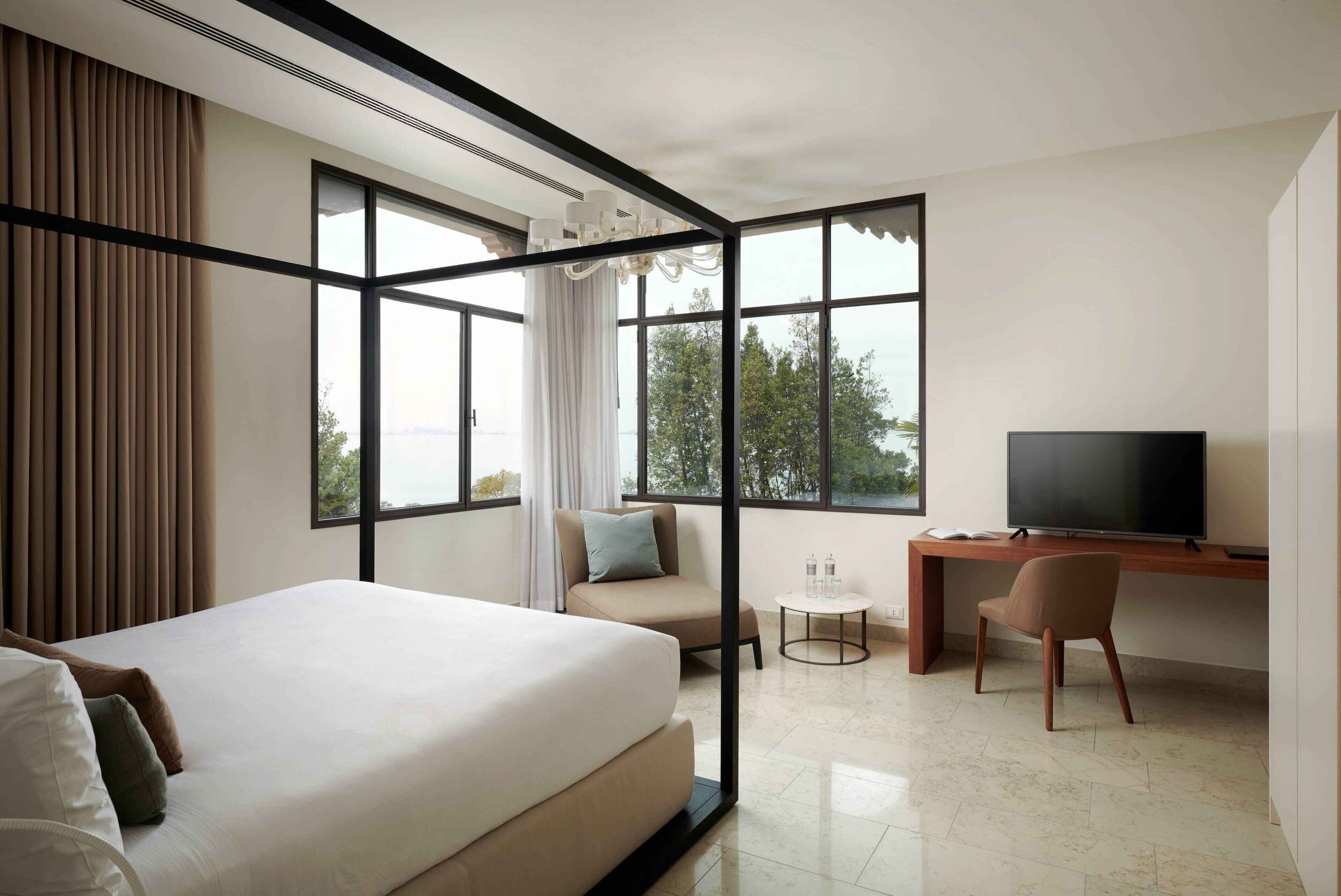 Een moderne hotelkamer met een groot bed, een bureau met een televisie en uitzicht op bomen door grote ramen.
