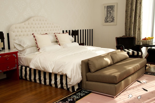 Un dormitorio ordenado con una cama grande, una mesita de noche roja, un sofá marrón y una pequeña mesa con una cesta de frutas.